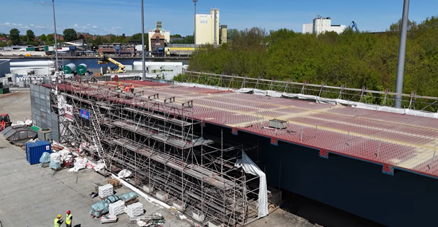 Luftaufnahme des rund 118 Meter langen Stahl-Brückenelements auf einer Hafenfläche, umgeben von Gerüstkonstruktionen, Baumaterialien und Arbeitskräften; im Hintergrund sind Industriegebäude und der Nord-Ostsee-Kanal zu sehen.